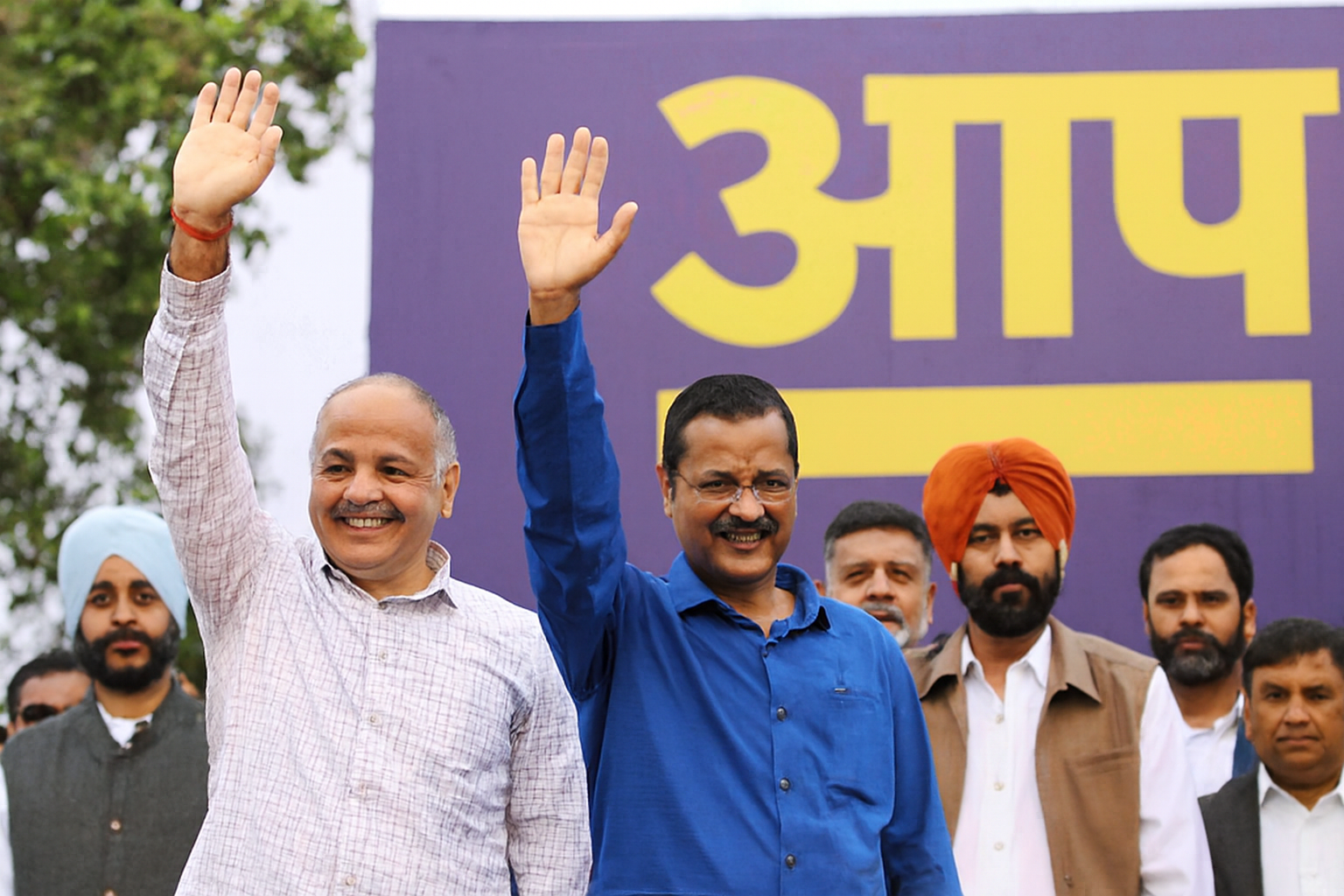  A balanced composition of leaders waving, men in turbans and casual attire behind them, bold yellow script in the background.